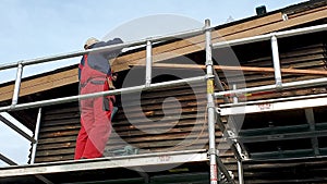 Worker on scaffolding installing new wooden planks on house roof eaves