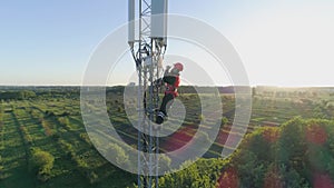 Worker serves cellular antenna with mobile phone in hand, aerial view of man in hard hat on background of city landscape