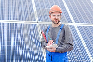 A worker at a solar power station in uniform looks somewhere and writes into a notebook holding waterpas.