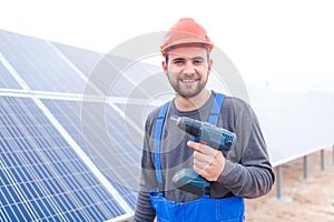 A worker at a solar station is smiling and holding a drill. Outdoors.