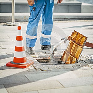 Worker standing next to open sewer manhole near traffic cone. Repair of sewerage and underground communications