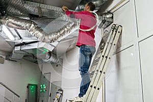 A worker standing on the stairs repairs the ventilation in the technical room of the residential complex. The installer works
