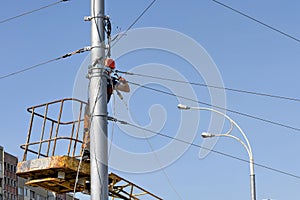 A worker tensions and secures a cable on a lamp post while standing in a lifting construction basket against a blue sky