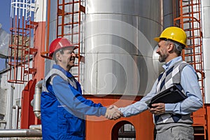 Worker in Uniform and Business Person Shaking Hands Against Power Plant or Oil Refinery Storage Tanks