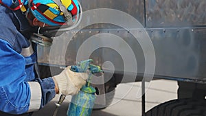 A worker in uniform and gloves paints a metal trailer with a compressor gun. Service for painting heavy machinery.