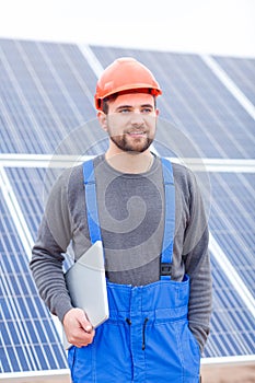 The worker in uniform stands near the solar battery panel and holds a laptop in his hands.
