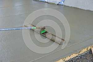 The worker was pouring concrete on the sidewalk using a long trowel on a wet cement surface, and he was leveling it as