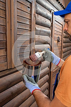 Worker wearing protective gloves and holding can with paint or varnish to protect wooden house exterior