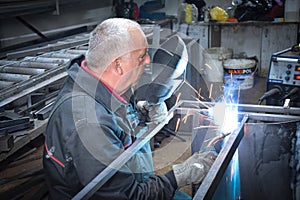 A Worker, welder in work clothes, construction gloves and a welding mask is welded with a welding machine metal product table,