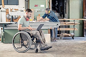 Worker in wheelchair with computer in a carpenter`s workshop with his colleagu