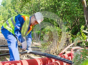 A worker in a white helmet and yellow vest, cuts a plastic pipe with blue scissors