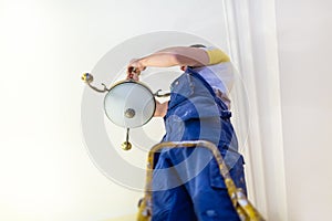 Worker in workwear is installing a chandelier and connects it to electricity on the ceiling from ladder in apartment i
