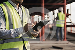 Worker in yellow vest, holding clipboard on construction site, overseeing progress photo