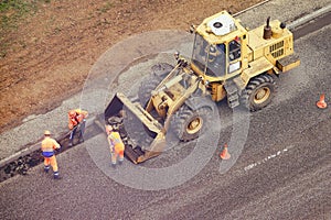 Workers with shovels in their hands remove asphalt from the road and putting it in the bucket of a bulldozer, top view