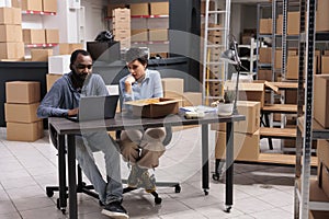 Workers sitting at desk table in storehouse checking shipping logistics on laptop computer