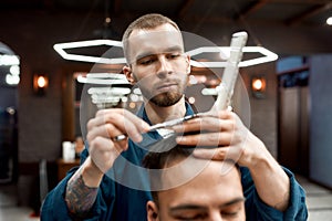 Working in barbershop. Close up of young professional barber with scissors and hair comb in hands making haircut for his