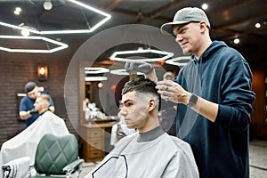 Working in barbershop. Professional barber in a baseball cap making hair styling for a young and handsome guy