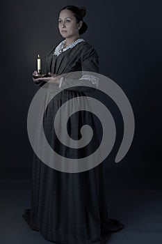 A working class Victorian woman holding a candle in the dark against a grey studio backdrop