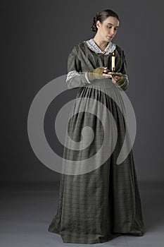 A working class Victorian woman wearing a dark green check bodice and skirt with an apron and holding a candle.