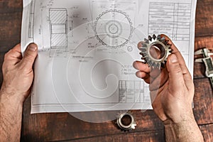 The working engineer holds a bevel gear in his hand against the background of a technical drawing