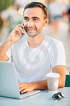 Working on fresh air. Handsome young man working on laptop and talking on the mobile phonewhile sitting at the wooden table outdoo