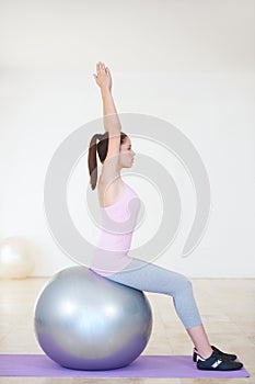 Working on her core. Profile shot of a young woman sitting on an exercise ball and stretching her arms above her head.