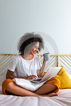 Working at home. African American young woman sitting on bed drinking hot coffee while typing on laptop. Vertical