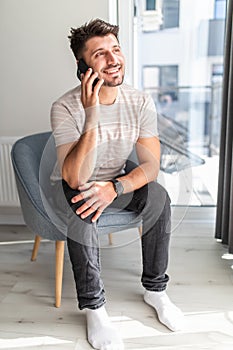 Working at home. Cheerful young handsome man working on laptop and talking on the mobile phone while sitting in big comfortable