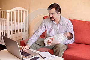 Working from home with kids: father in grey shirt sitting on sofa, holding in one hand infant baby and typing on laptop keyboard