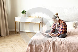 Working from home. Ordinary caucasian woman in casual wear and headset sits on the bed and using laptop, selective focus