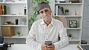 Working with joy, a confident young hispanic man enjoys his work at the office, handsomely typing messages on his smartphone while