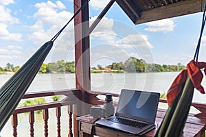 Working with laptop on wooden balcony in tourist resort with hanging hammock on the Mekong River, Laos. Concept of millenials