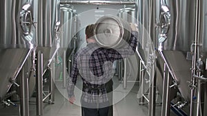 A working male brewer carries a keg filled with beer as he passes beer tanks. back view
