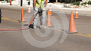 Working man uses a jackhammer on the asphalt of a city street next to the orange safety warning cones, as part of a utility