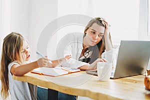Working mom freelancer on laptop with little daughter drawing on table at the home