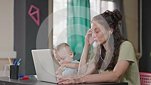 working mom and her little child in apartment, woman works online by laptop, boy is playing near