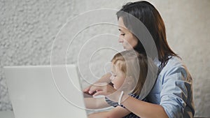 Working mother with baby at table. Busy woman working on laptop with little girl smiles and laughs on hands.