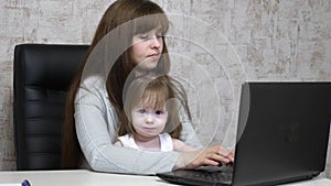 Working mother with her little daughter at the table. Busy woman working on laptop with baby on hands. Working mom with
