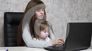 Working mother with her little daughter at the table. Busy woman working on laptop with baby on hands. Working mom with