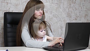 Working mother with her little daughter at the table. Busy woman working on laptop with baby on hands. Working mom with