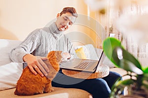 Working online from home with cat using computer. Man holding laptop sitting on couch by ginger pet looking at screen.