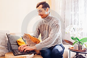 Working online from home with pet using computer. Man typing on laptop with ginger cat looking at screen.