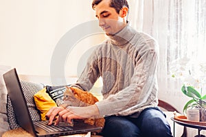 Working online from home with pet using computer. Man typing on laptop keyboard with ginger cat looking at screen.