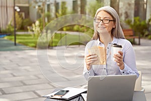 Working online. Portrait of a happy mature business woman wearing eyeglasses having a lunch, drinking coffee and eating