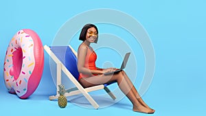 Working online from tropical paradise. Black woman in bikini sitting in lounge chair, using laptop on blue background