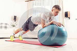 Working out at home by young man holding a plank position using a blue fit ball and red mat inside a living room