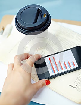 Working outside the office. Hand holding smartphone with bar chart on screen with disposable cup, pen, newspaper and