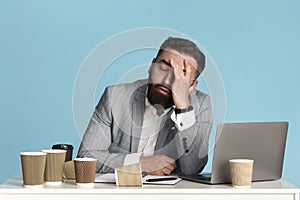 Working overtime. Exhausted businessman sitting at desk with laptop and empty coffee cups against blue background