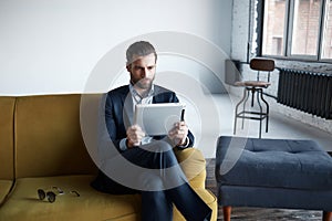 Working...Successful and fashionable businessman is using a tablet while sitting on sofa at modern office