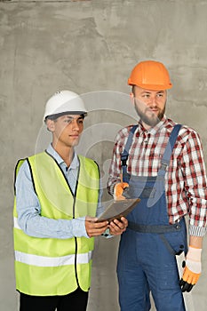 Working in team. Two young and cheerful builders in protective helmets are using digital tablet and working while